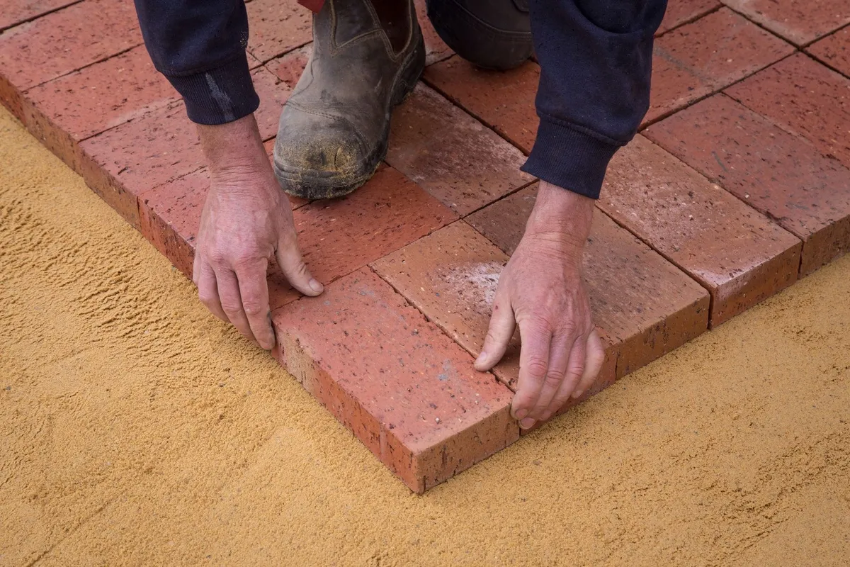 A worker in uniform laying bricks in a square pattern on sandy ground for landscaping.